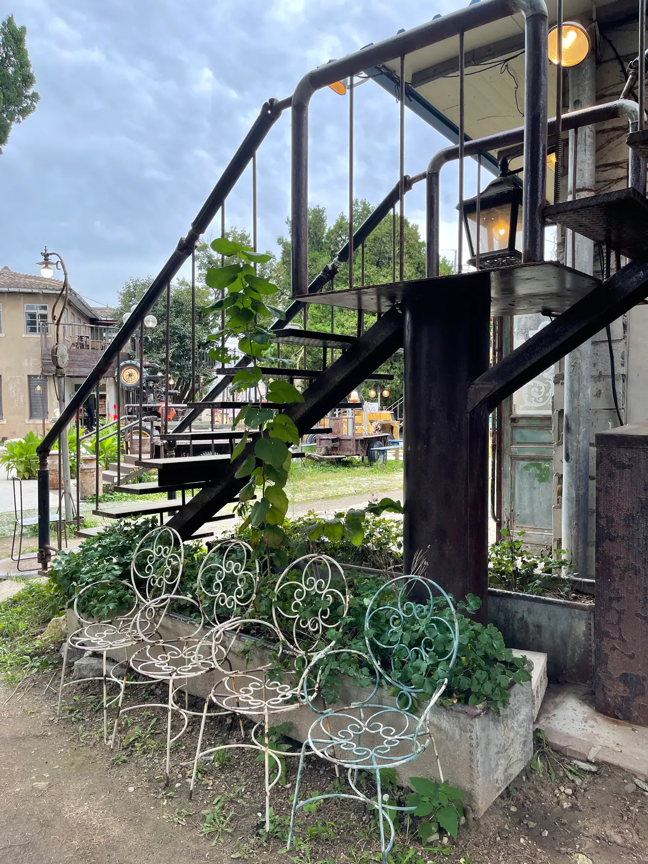 White wrought-iron chairs stand in an overgrown planter under a black steel staircase.