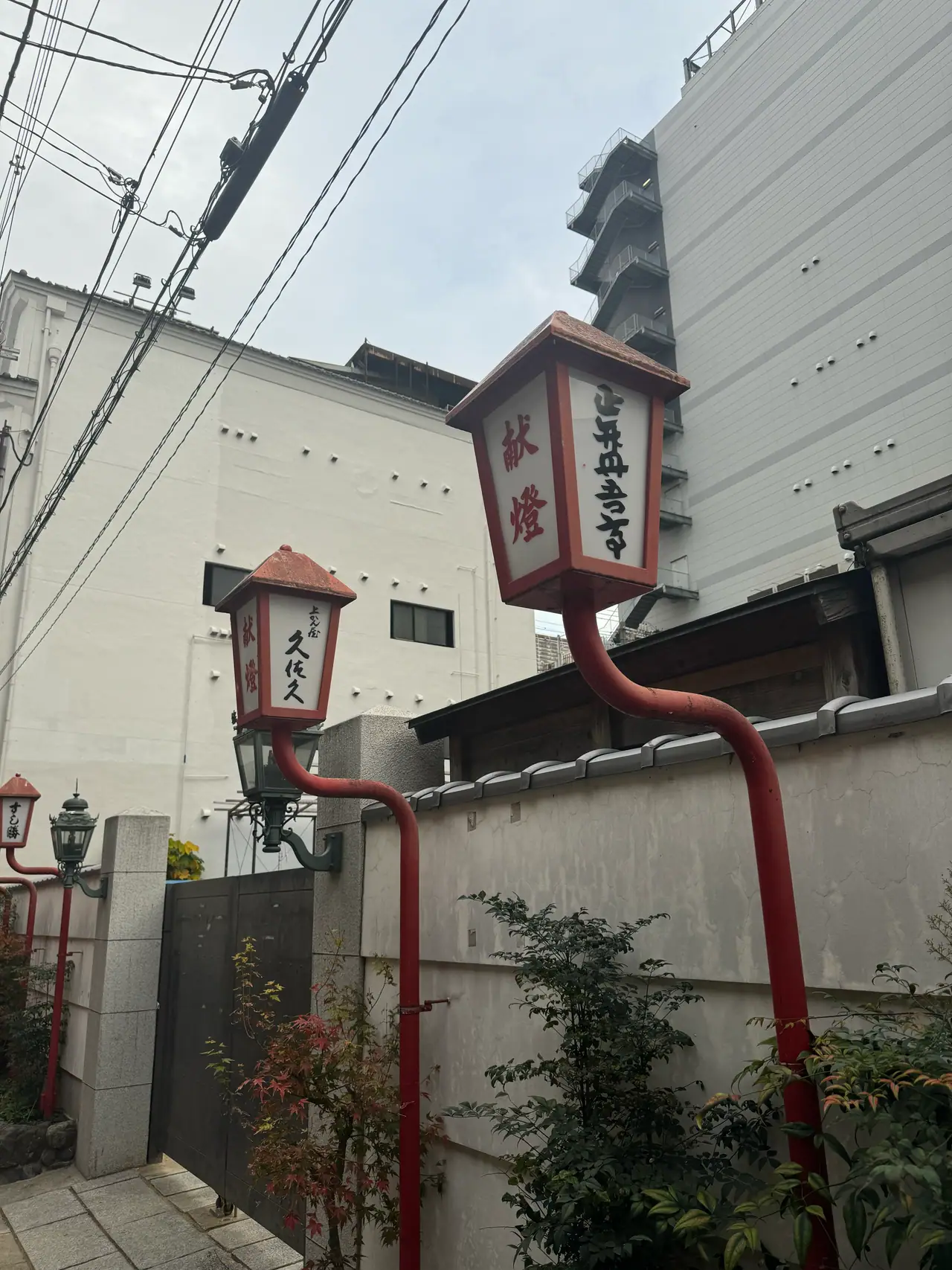 Two red lantern-topped street lamps stand against a white plaster wall under a tangle of power lines.