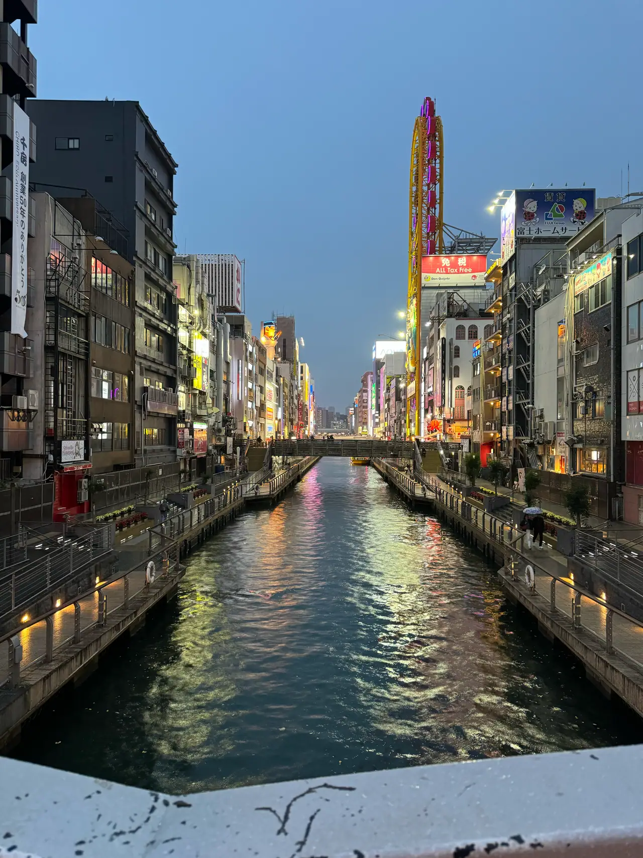 Neon signs line both banks of a canal at dusk, their light doubled on the still water.
