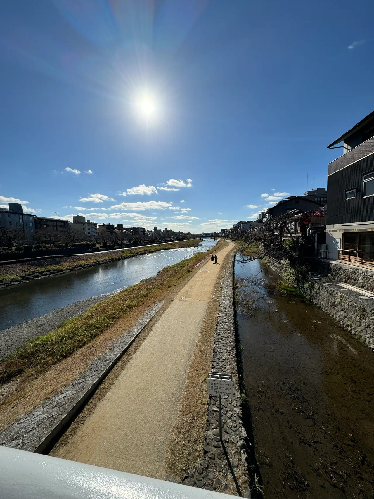 The Kamo river runs straight toward the horizon under a white sun, its walkways empty on both banks.