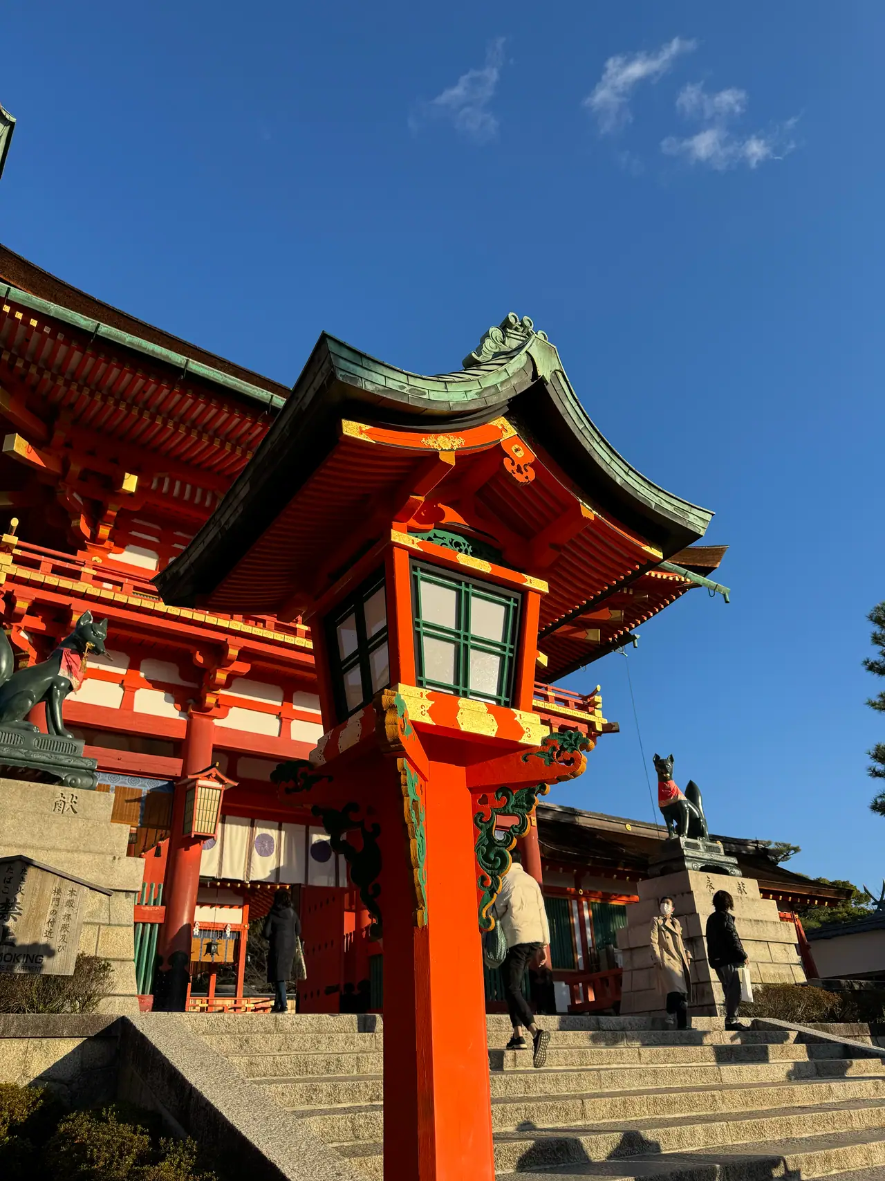 A vermilion shrine gate with green copper eaves and a hanging lantern rises against a clear sky.