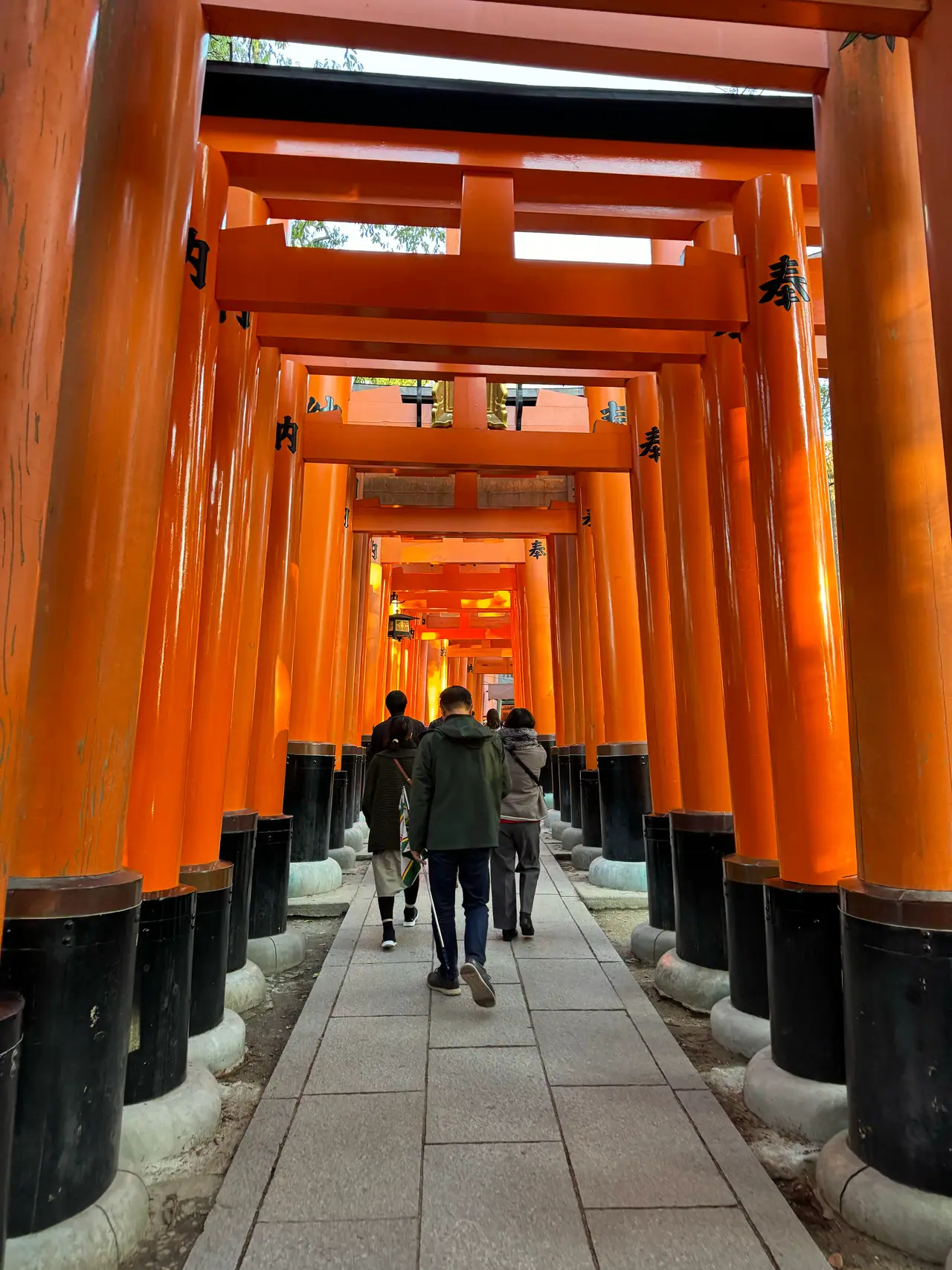 A tunnel of vermilion torii recedes to a bright point, three people walking through it.