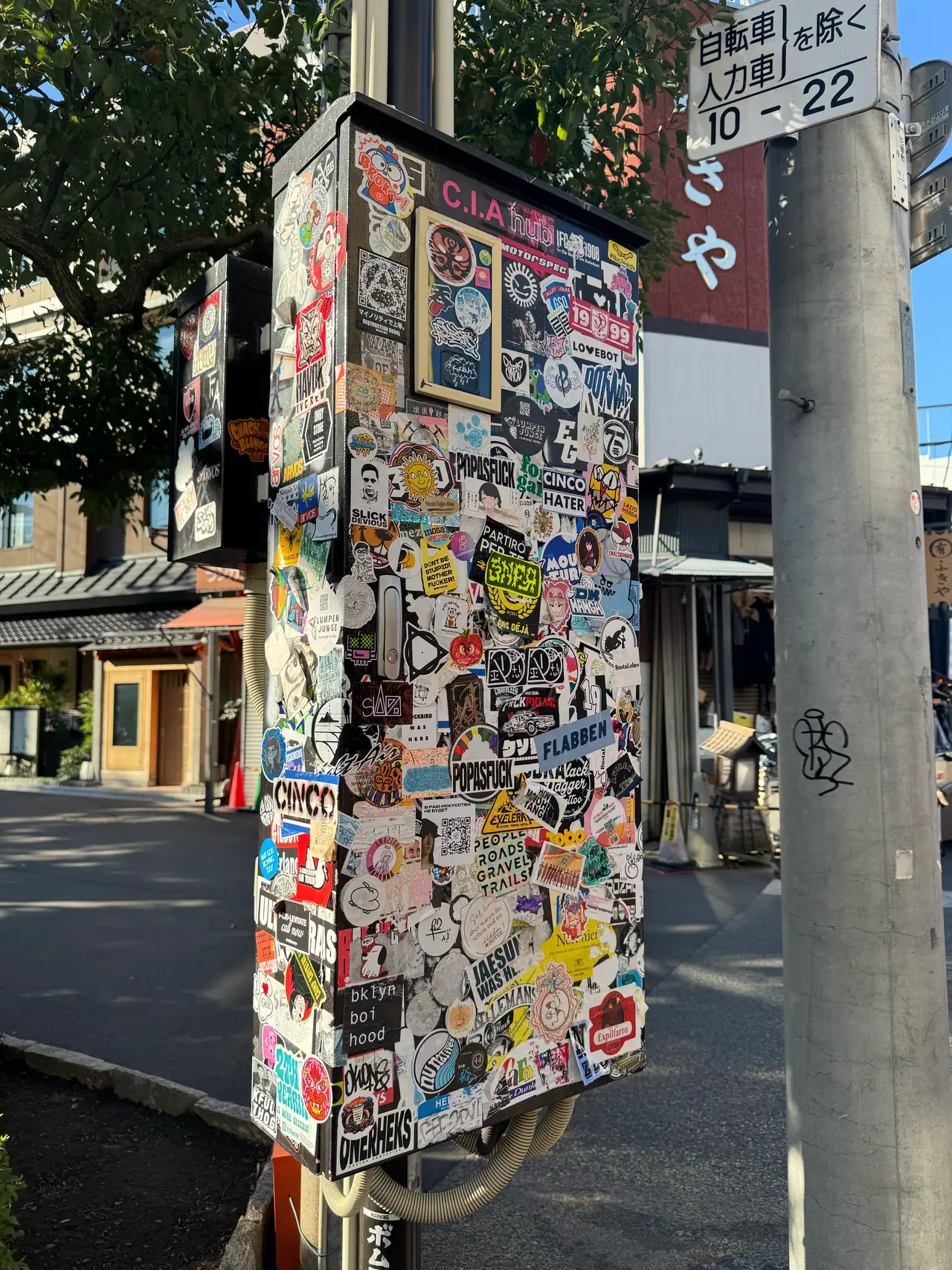 A utility cabinet is covered end to end in overlapping stickers beside a bare concrete pole.