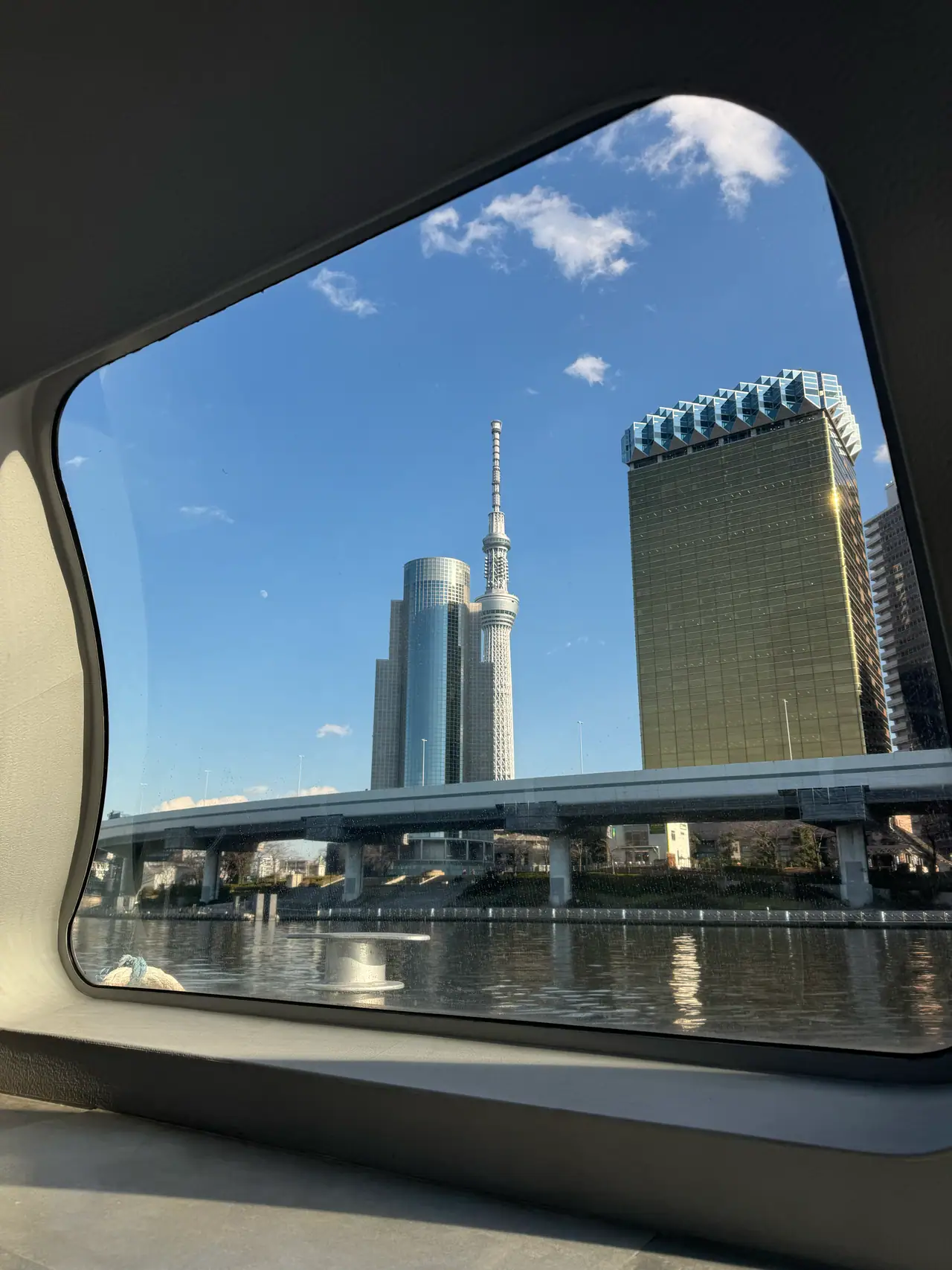 A rounded water-bus window frames the Skytree and a gold-clad tower across the river.
