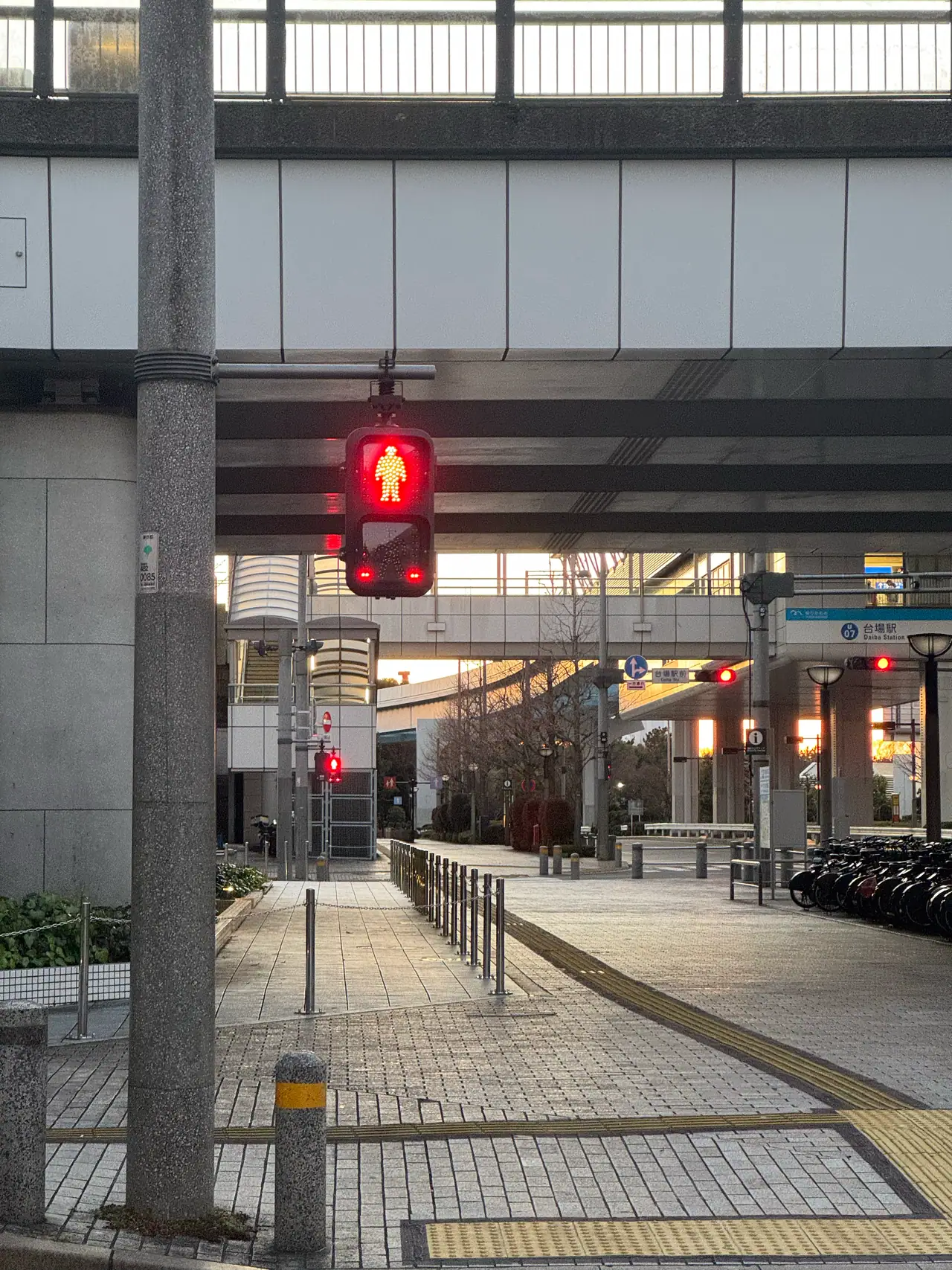 A red pedestrian signal glows under an elevated deck above an empty paved walkway.