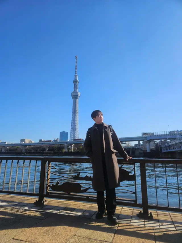 A person leans against a riverside railing with the Skytree rising across the water behind them.