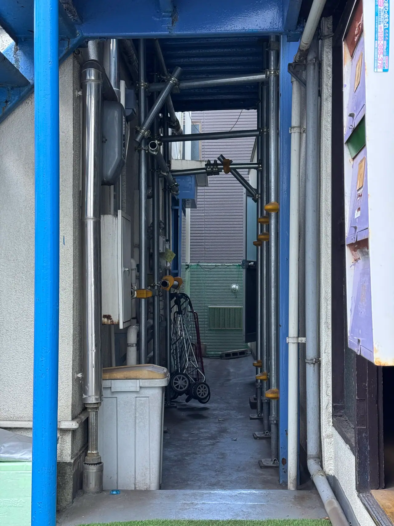 A narrow service passage under a blue roof runs between two buildings toward daylight.