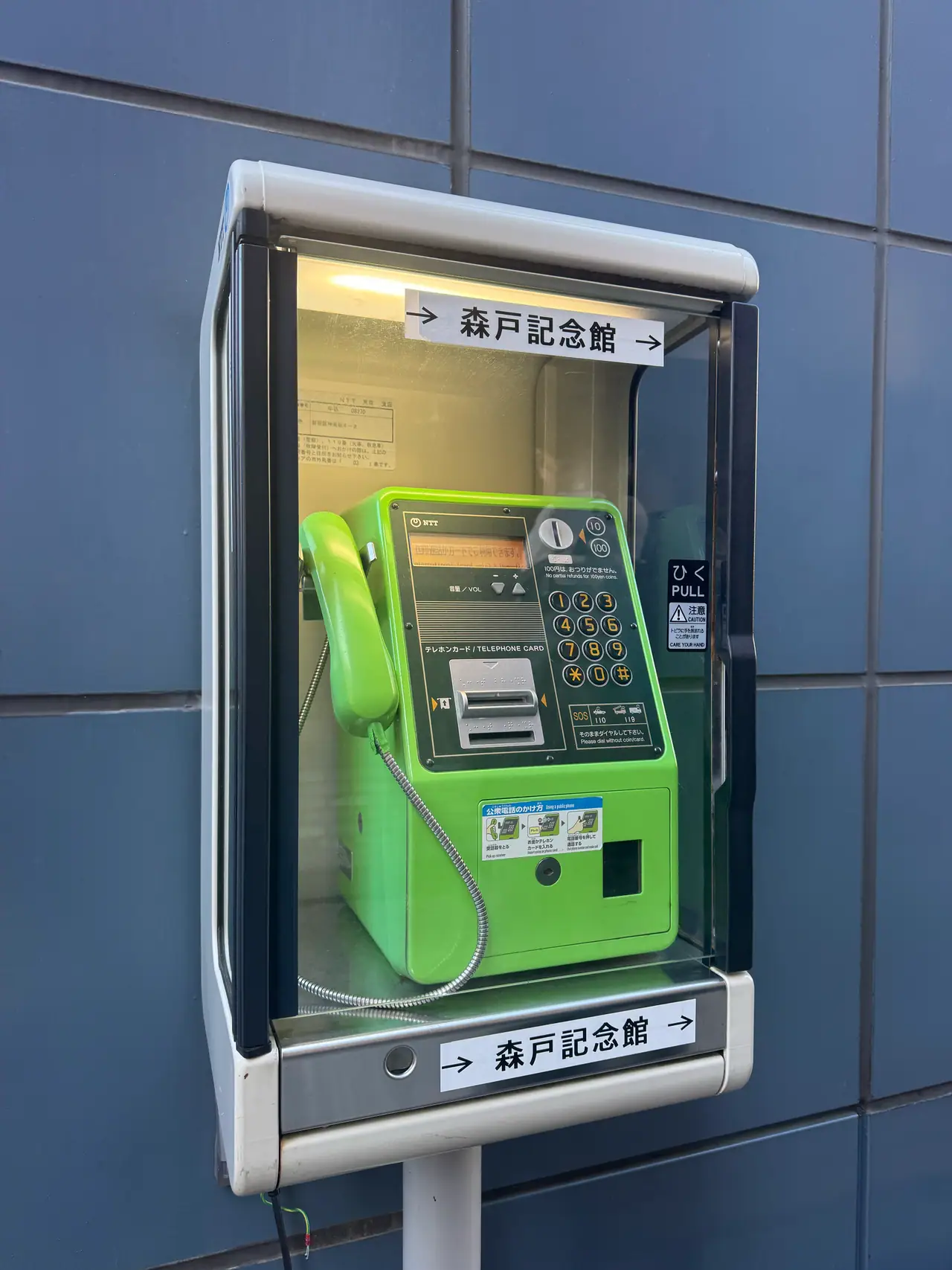 A green payphone sits in a metal booth against a tiled wall, direction signs above and below it.