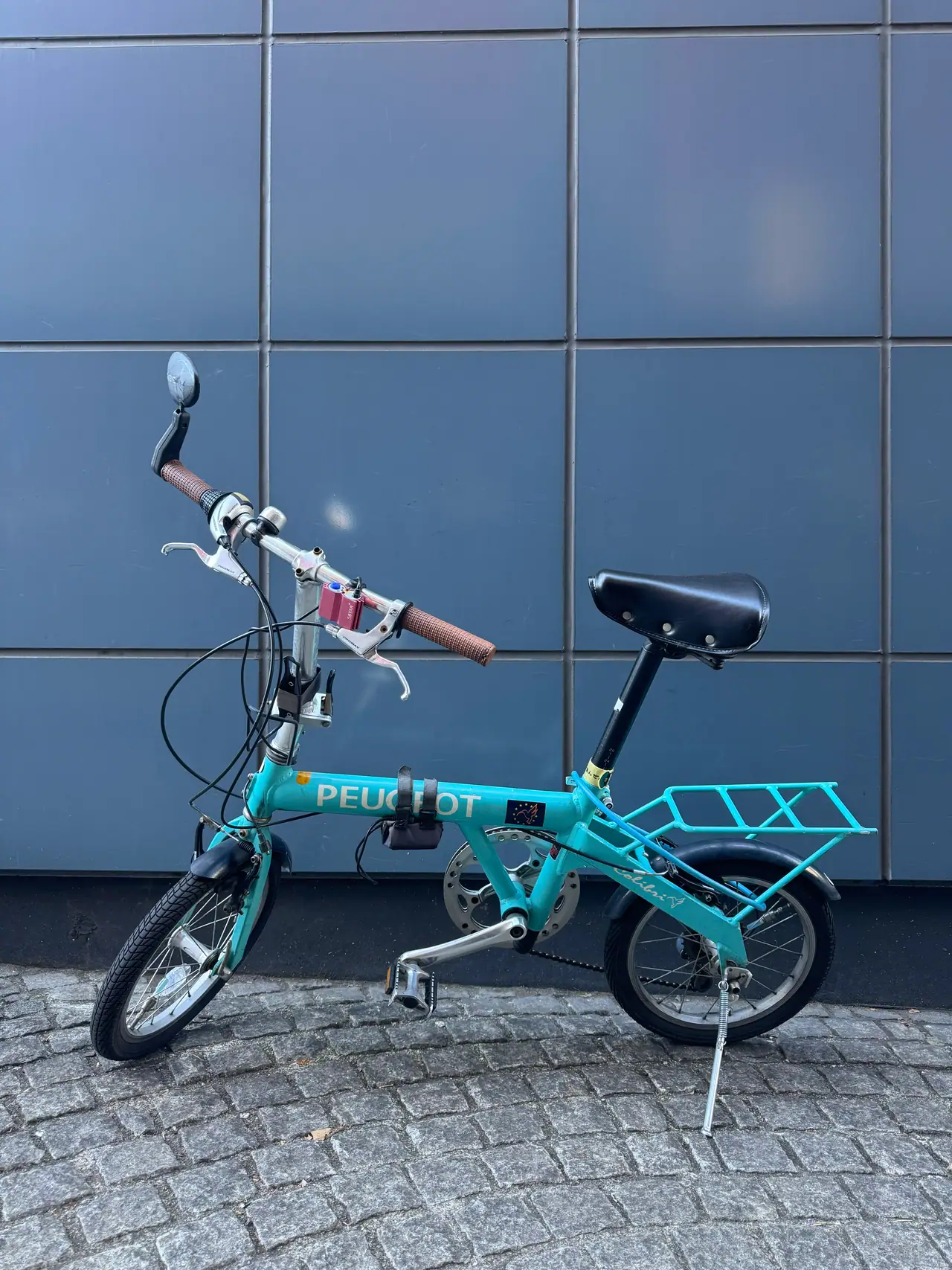 A teal folding bicycle stands on its kickstand against a grid of pale wall tiles.