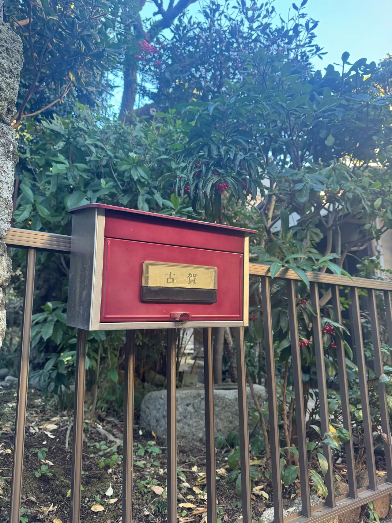 A small red wooden box for spent shrine charms hangs on an iron fence among winter foliage.