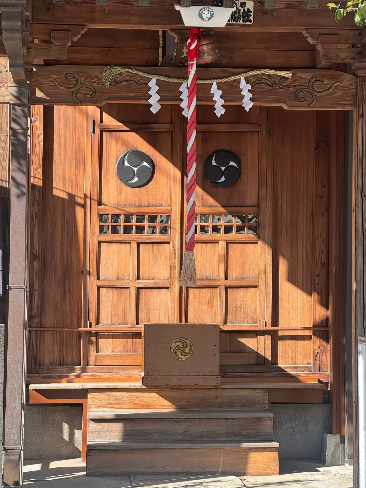 Sunlight falls across wooden shrine doors marked with two dark crests, a rope and paper streamers above.