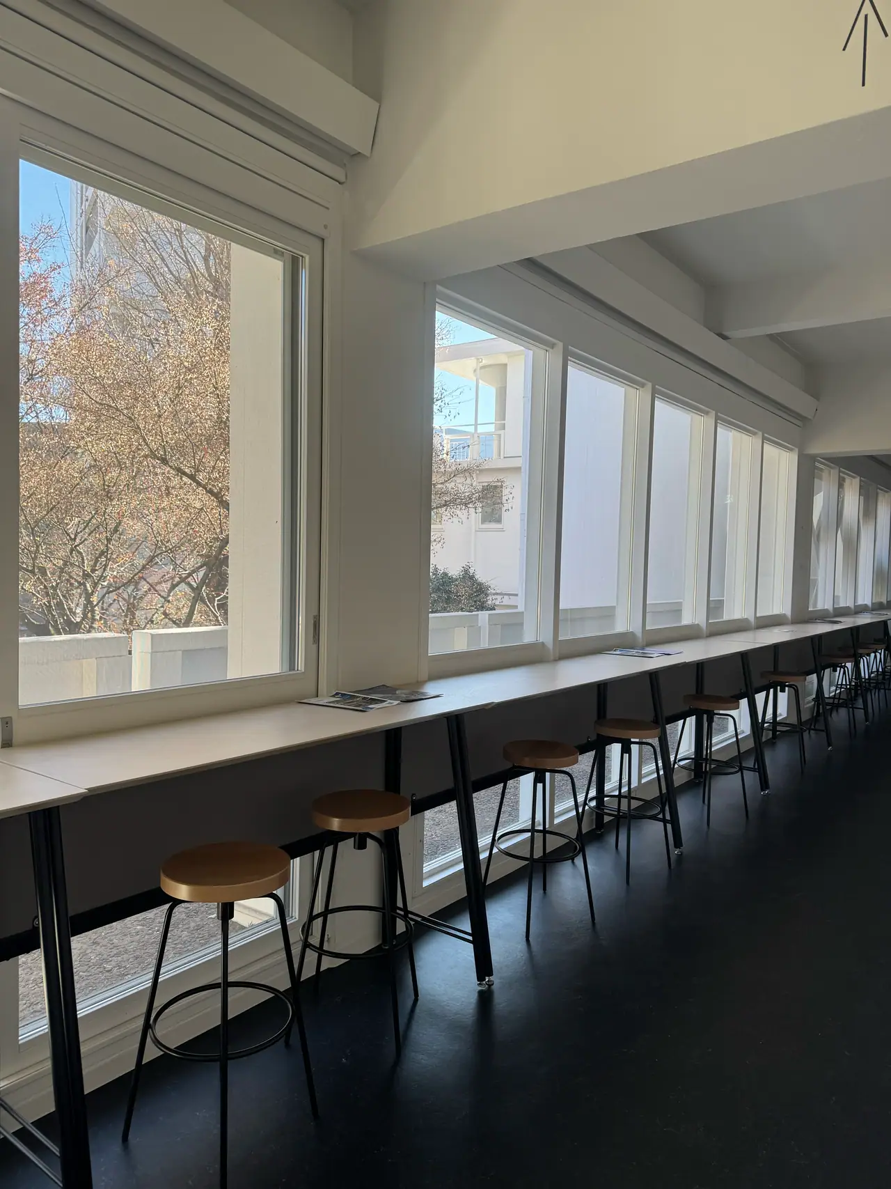 A long wooden counter and stools run beside a wall of windows in a white room.