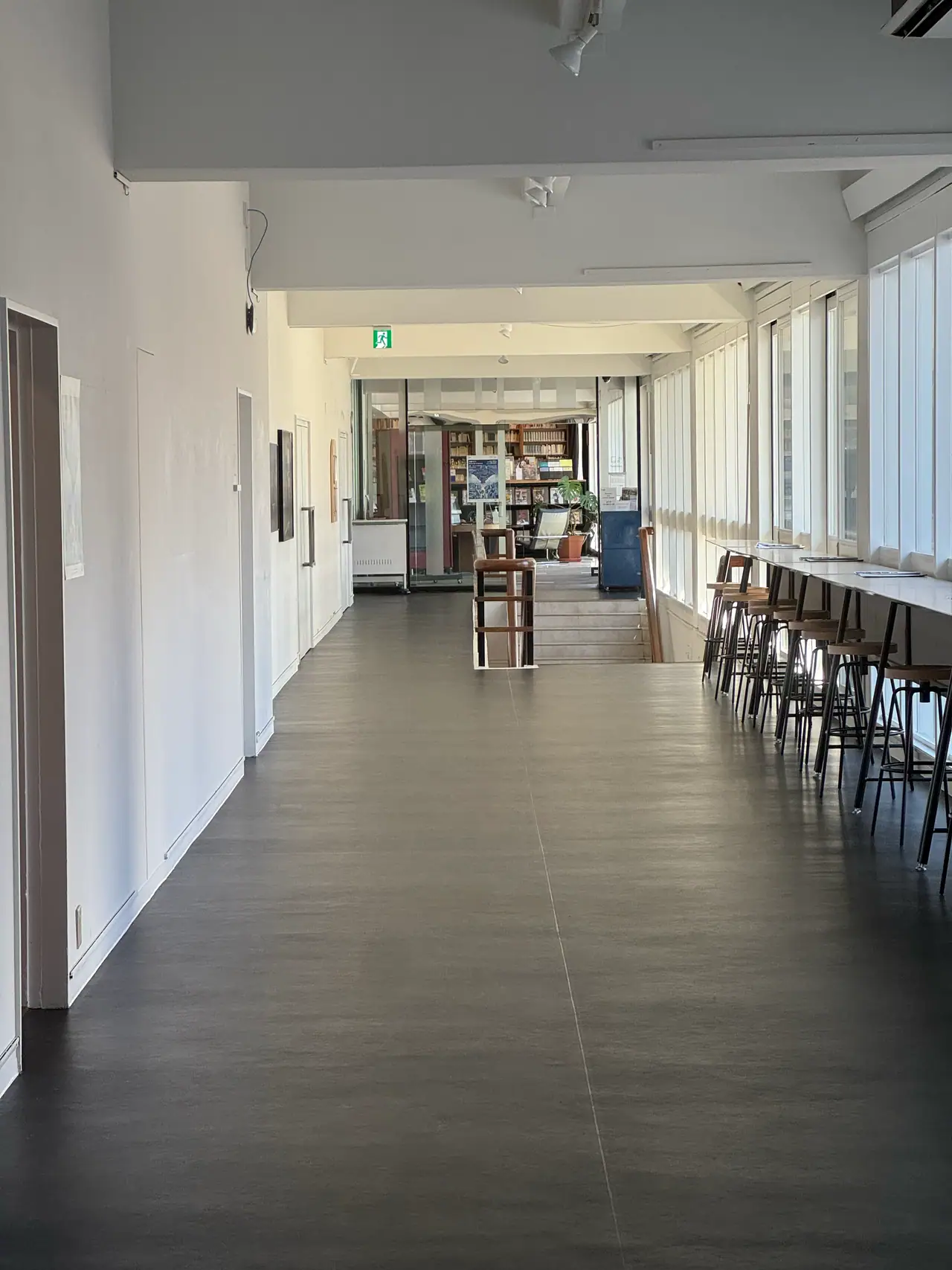 A white corridor with a dark floor runs past a row of stools toward a lit room at the far end.