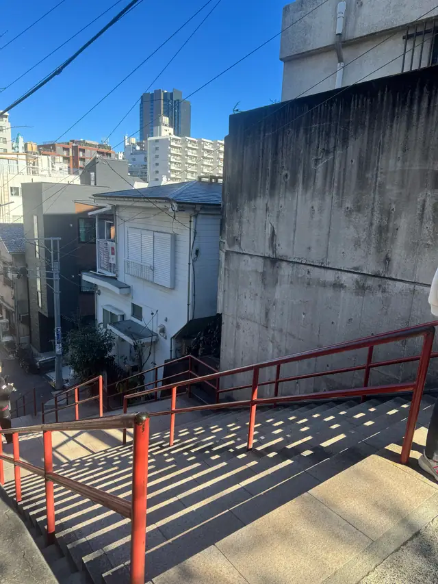 A red handrail throws a ladder of shadows across a paved slope below a concrete retaining wall.