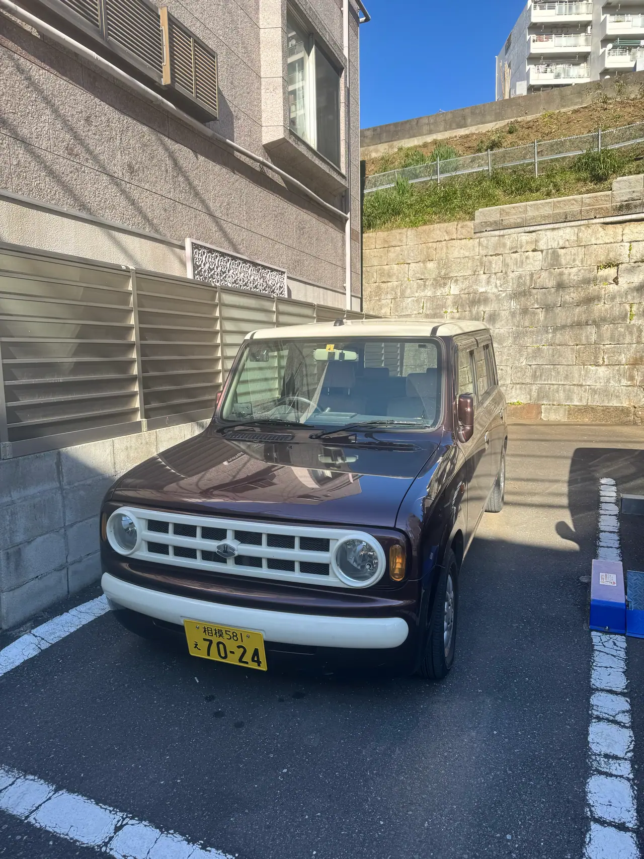 A dark red kei van is parked against a concrete block retaining wall on a bright street.