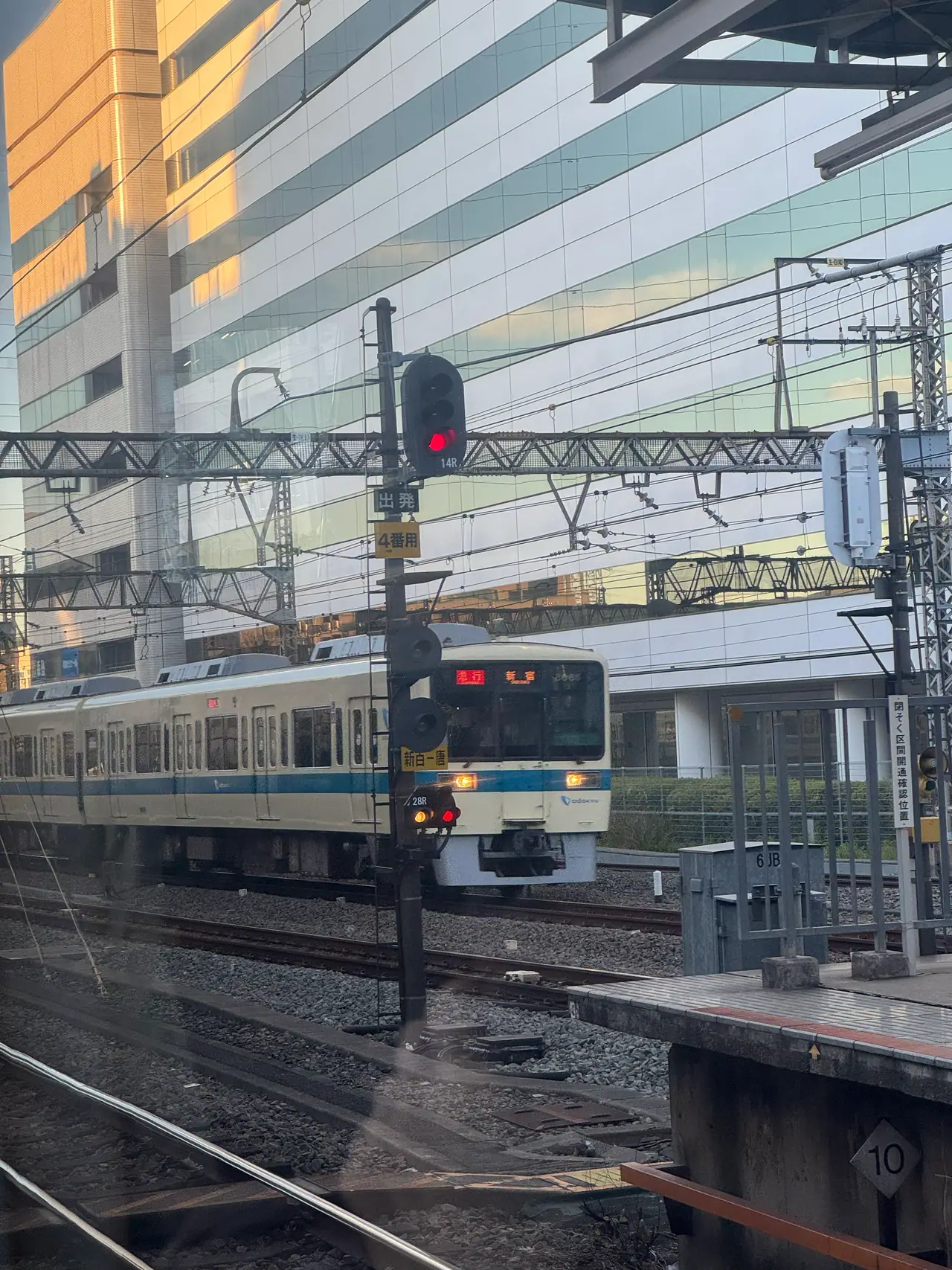 A commuter train waits beside a platform, seen through a window laced with overhead wires and signals.