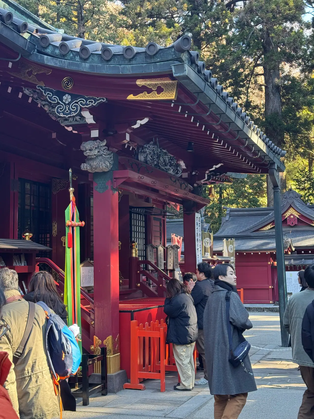Visitors gather in front of vermilion shrine buildings beneath tall trees.