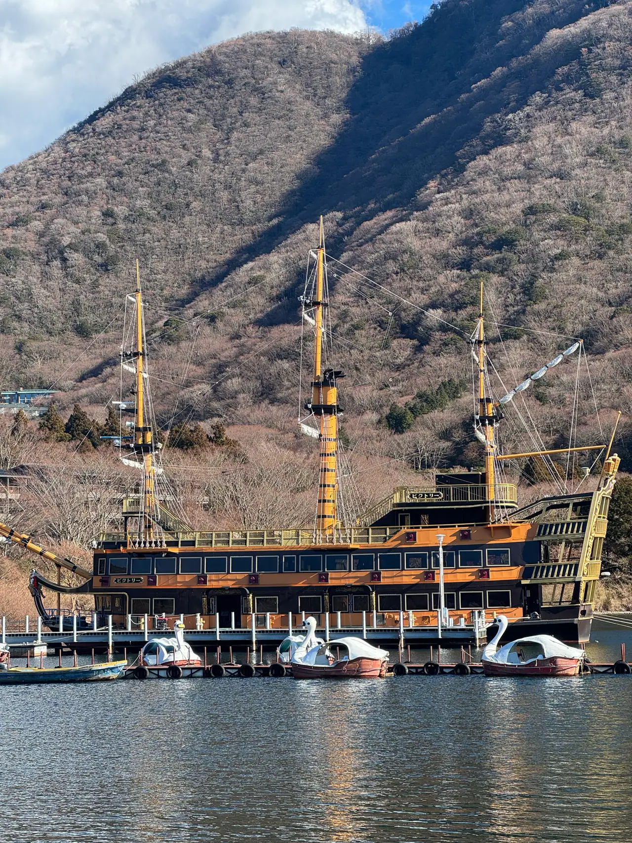 White swan pedal boats float in front of a moored galleon beneath a bare mountain.