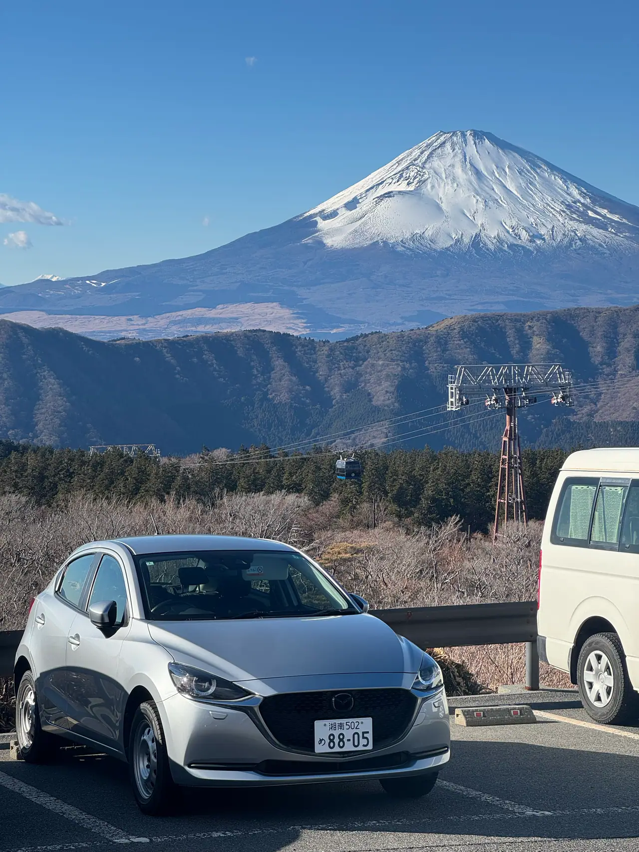 A silver hatchback is parked at a roadside with Mount Fuji rising beyond the ridge.
