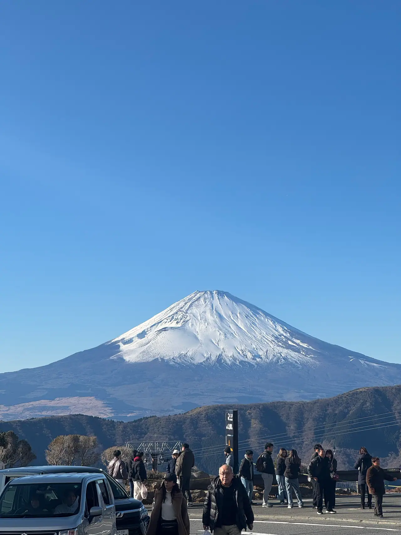 A crowd stands along a roadside barrier looking toward Mount Fuji across a wide valley.