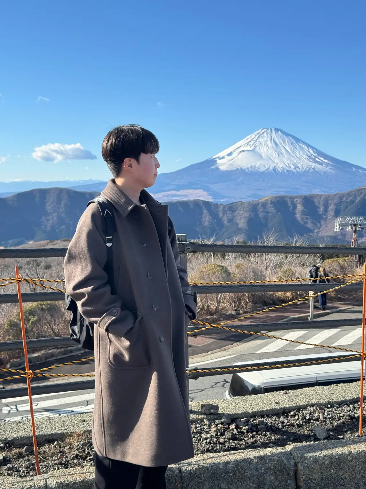A person in a long coat stands at a barrier with Mount Fuji in the distance behind them.