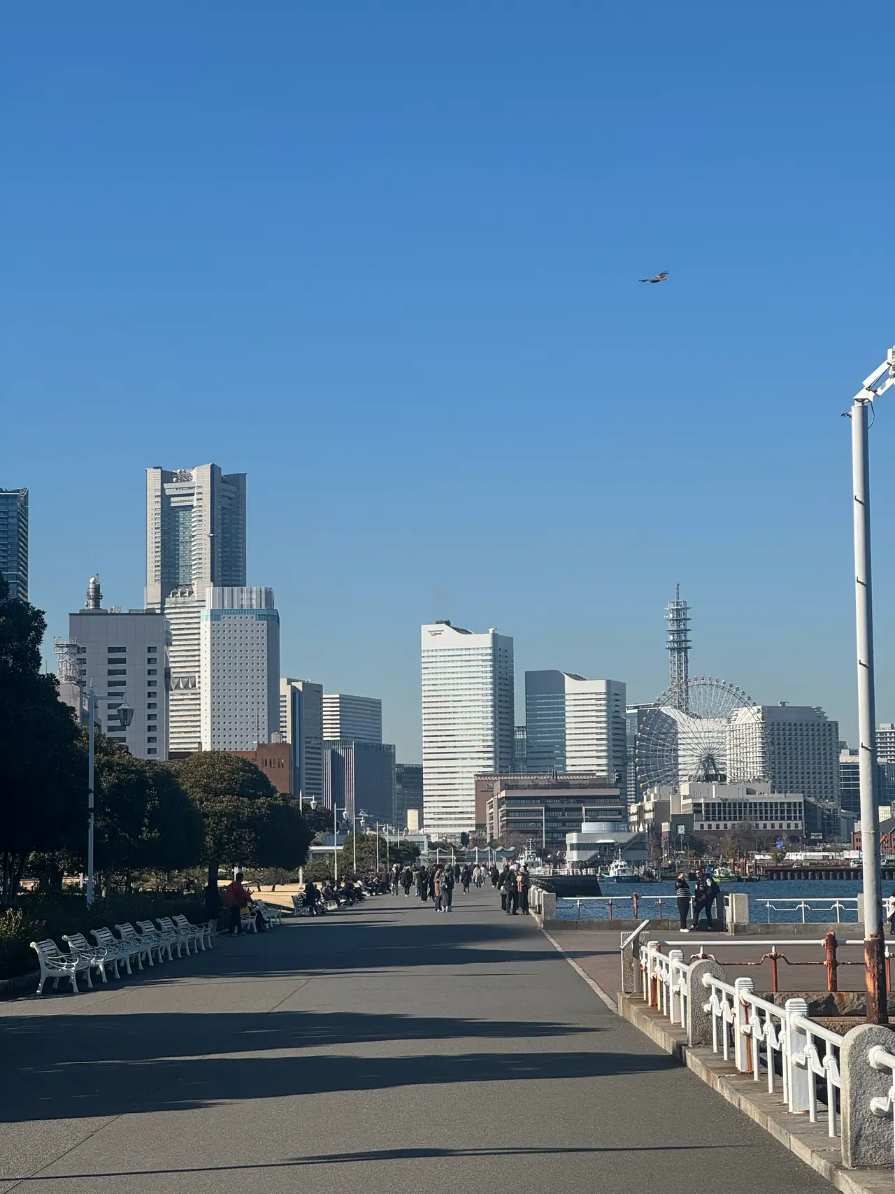 A wide waterfront promenade runs toward the Minato Mirai skyline, a plane crossing the sky above.